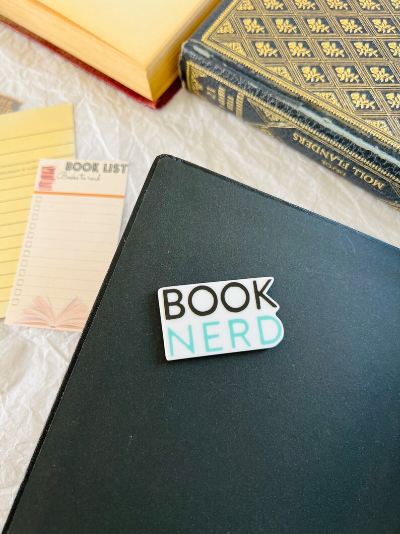 Op de afbeelding: Een zwart notitieboek met een witte sticker waarop "BOOK NERD" in zwart en blauw staat.