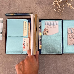Könnte beinhalten: Ein dunkelbraunes Leder-Reisenotizbuch mit metallisch hellblauen Innentaschen. Es enthält einen Notizblock, eine To-Do-Listen-Karte und eine Karte mit dem Text 'All you need is love a passport'. Eine separate Karte trägt die Aufschrift 'YOU TASTE LIKE SUNSHINE'.