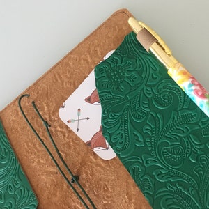 K&ouml;nnte beinhalten: Ein handgefertigtes Lederjournal mit gr&uuml;n gepr&auml;gtem Einband und einer Karte mit Fuchsbildern. Ein goldener Stift mit farbenfrohem Design liegt auf dem Journal. Das Journal ist ge&ouml;ffnet und zeigt das Innere und eine Karte mit Fuchsbildern.