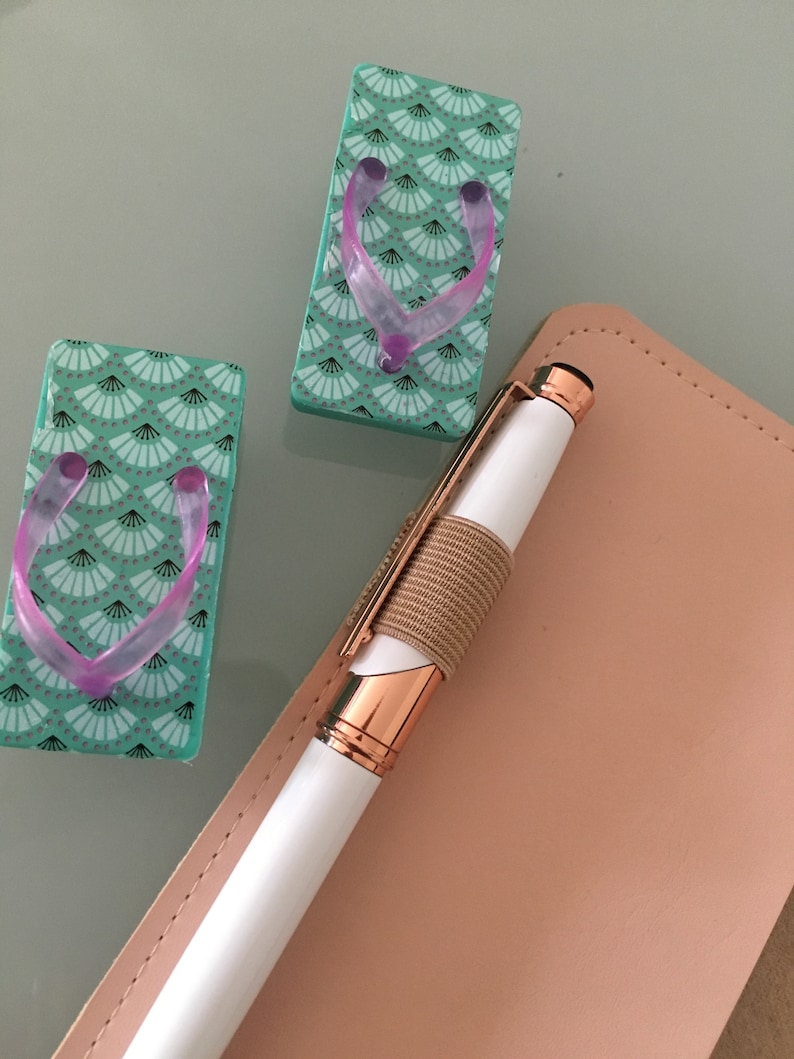 K&ouml;nnte beinhalten: Zwei dekorative Miniatur-Geta-Sandalen mit einem t&uuml;rkisfarbenen F&auml;chermuster und rosa Riemen. Ein wei&szlig;er Stift mit ros&eacute;goldenen Akzenten und einem beigefarbenen Gummiband liegt auf einem rosa Leder-Notizbuch.