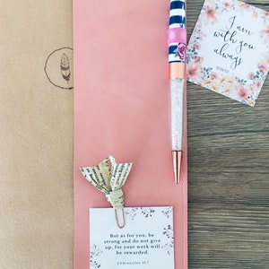 K&ouml;nnte beinhalten: Ein rosa Lederjournal mit einem Stift, einer Papierblume und einer Karte. Der Stift hat einen Kristallk&ouml;rper, ros&eacute;goldene Akzente und ein gestreiftes Design. Auf der Karte steht "Ich bin immer bei dir." Eine andere Karte lautet "Aber f&uuml;r dich, sei stark..."