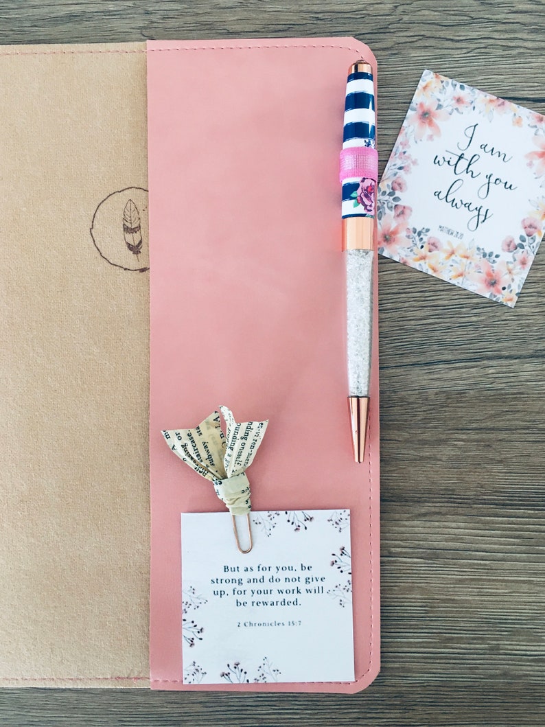 K&ouml;nnte beinhalten: Ein rosa Lederjournal mit einem Stift, einer Papierblume und einer Karte. Der Stift hat einen Kristallk&ouml;rper, ros&eacute;goldene Akzente und ein gestreiftes Design. Auf der Karte steht "Ich bin immer bei dir." Eine andere Karte lautet "Aber f&uuml;r dich, sei stark..."