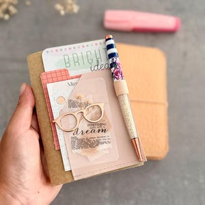 Könnte beinhalten: Ein beigefarbenes Notizbuch mit einer Tasche, die eine Karte mit der Aufschrift BRIGHT ideas und eine Karte mit einem roségoldenen Brillensymbol und dem Text EVERYTHING starts with a dream enthält. Ein Stift mit blau-weißem Blumenmuster ist befestigt.
