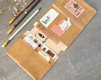 May include: A tan leather journal with multiple pockets, open to display cards, stickers, and a clear pocket with the text "Enjoy the small things." Three pencils and a sprig of dried flowers are next to the journal. The word "love" is printed on a white card.