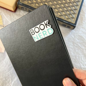 Op de afbeelding: Een zwart lederen gebonden dagboek met een witte sticker waarop in zwarte en turquoise letters "BOOK NERD" staat.
