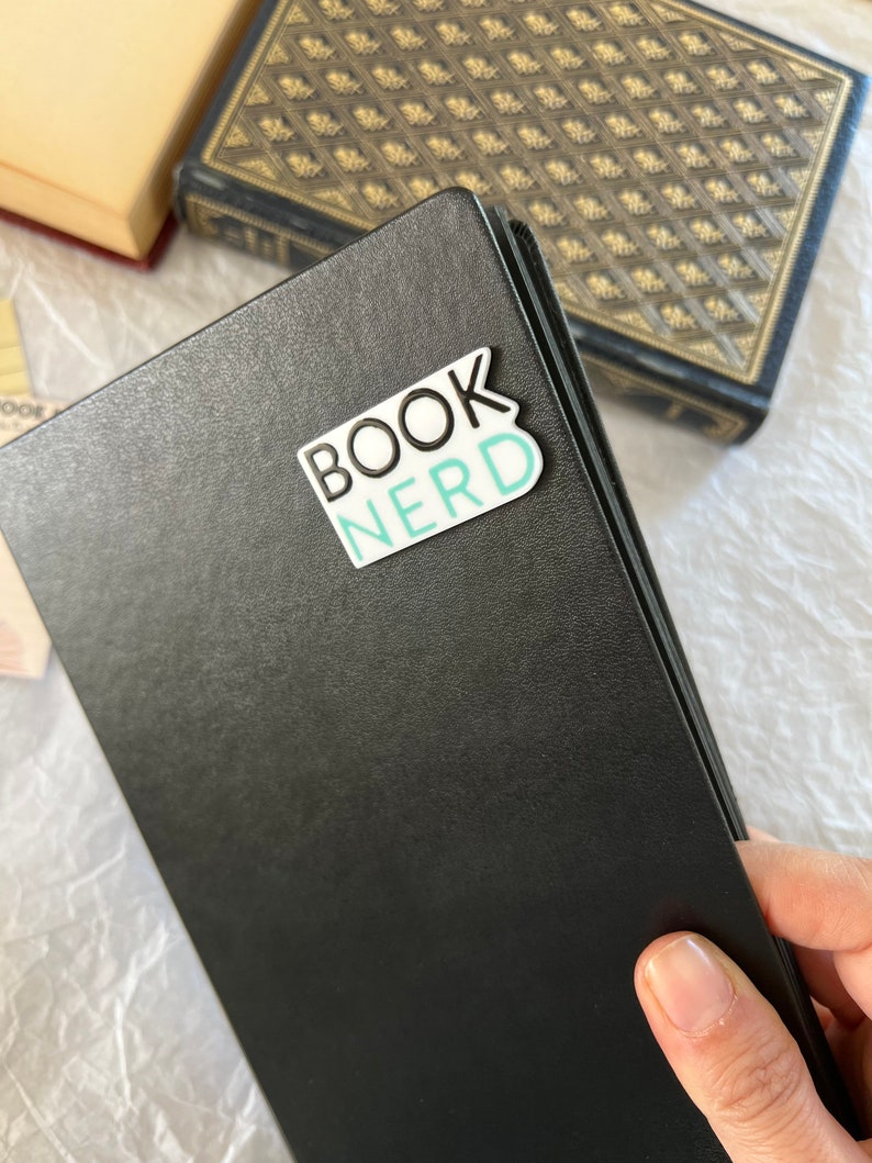 Op de afbeelding: Een zwart lederen gebonden dagboek met een witte sticker waarop in zwarte en turquoise letters "BOOK NERD" staat.