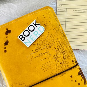 Op de afbeelding: Een geel lederen gebonden dagboek met een bruine elastische band sluiting. Het dagboek heeft een sticker aan de voorkant met de tekst "Book Nerd" in zwarte en blauwe letters.