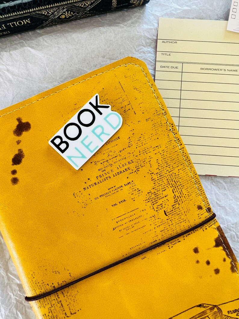 Op de afbeelding: Een geel lederen gebonden dagboek met een bruine elastische band sluiting. Het dagboek heeft een sticker aan de voorkant met de tekst "Book Nerd" in zwarte en blauwe letters.