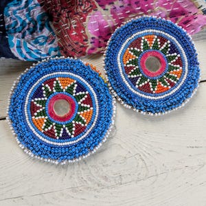 May include: Two circular patches featuring intricate beadwork in shades of blue, orange, green, white, and red. Each patch has a central circular mirror surrounded by a star-shaped pattern of beads, with a thick blue beaded border around the edge.