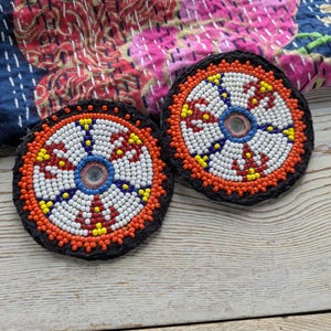 Puede incluir: Dos posavasos redondos con cuentas y borde negro. Los posavasos presentan un espejo central rodeado de cuentas blancas, rojas, azules y amarillas. El fondo incluye un textil estampado colorido y una superficie de madera clara.