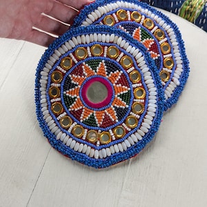 Peut inclure: Trois dessous de verre ronds et perlés avec des motifs complexes. Les dessous de verre présentent un miroir central entouré d'un motif en étoile rouge, orange et vert. Des accents dorés et des perles blanches bordent le motif, avec un bord perlé bleu.