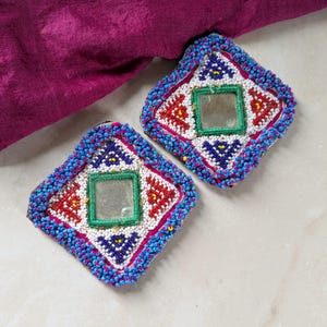 May include: Two square, beaded decorative patches with a central mirror. The patches feature red, white, blue, and yellow beadwork in a geometric pattern. The edges are outlined with blue beads, and a magenta fabric is visible in the background.