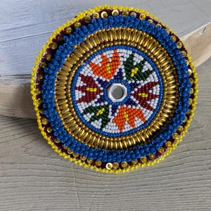 May include: A circular beaded patch featuring a central small mirror surrounded by a radial pattern of white, orange, green, and dark red beads. The design includes concentric rings of metallic gold tube beads, blue seed beads, and a yellow beaded edge.