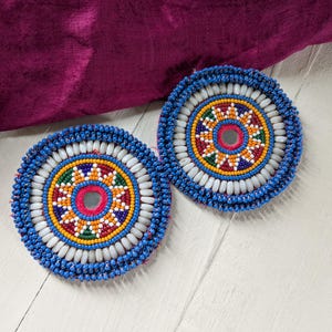 May include: Two round, beaded coasters with intricate designs. The coasters feature a central mirror surrounded by colorful beads in red, yellow, green, and blue. White beads and a blue border complete the design. The coasters rest on a white surface.