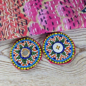 May include: Two round, beaded embellishments with a central mirror and button. The beads are arranged in concentric circles of blue, green, orange, and white, set on a pink felt base. A patterned textile is in the background.