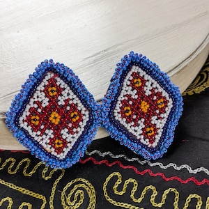 May include: A pair of diamond-shaped earrings featuring intricate beadwork in blue, white, red, and yellow. The design consists of a central yellow bead surrounded by red and white geometric patterns, all enclosed by a thick border of blue beads.