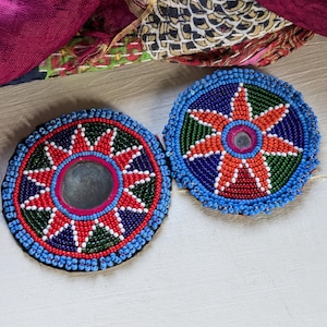May include: Two circular coasters featuring intricate beadwork in vibrant shades of blue, red, green, and white. Each coaster displays a central star-shaped pattern surrounding a small, reflective circular mirror element at the very center.