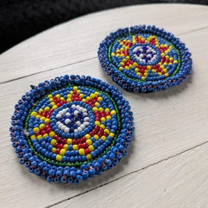 May include: Two round, beaded coasters. The coasters feature a vibrant design with blue, red, yellow, and white beads. The outer edge is a deep blue, with concentric circles of color radiating inward. The coasters are placed on a white wooden surface.