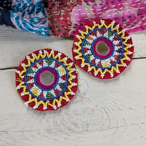 Puede incluir: Dos parches de tela circulares con un espejo central rodeado por anillos concéntricos de cuentas de vidrio multicolores en rojo, amarillo, azul, verde y blanco. El borde exterior de cada parche tiene un patrón en zigzag de cuentas amarillas.