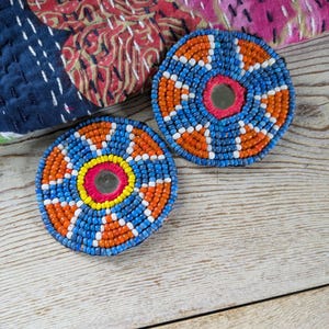 May include: Two round beaded coasters. The coasters feature a central mirror surrounded by a ring of pink beads, then a ring of yellow beads, and then a pattern of blue, orange, and white beads. The coasters are on a light wood surface.