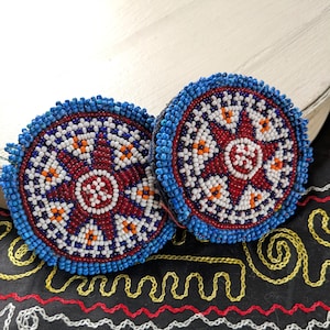 May include: Two circular earrings featuring intricate beadwork in a starburst pattern. The design incorporates small blue, white, red, and orange glass beads arranged in concentric circles and geometric shapes, with a thick blue beaded border.