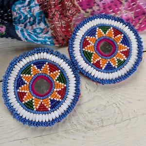May include: Two circular coasters featuring intricate beadwork in a sunburst pattern. Each coaster has a central mirror surrounded by orange, red, green, and blue beads, finished with a thick border of white and blue beads on the outer edge.