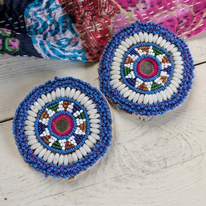 May include: Two circular coasters featuring intricate beadwork in blue, white, green, and brown. Each coaster has a central reflective mirror element surrounded by concentric rings of small glass beads and larger oval white beads.