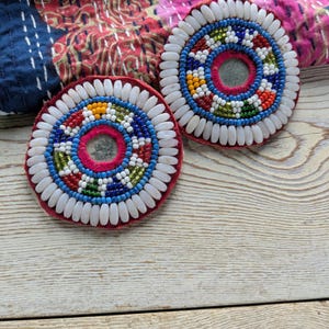 Puede incluir: Dos posavasos redondos con cuentas y respaldo de terciopelo rojo. Los posavasos presentan un espejo central rodeado de círculos concéntricos de cuentas de colores y cuentas blancas de forma ovalada. Los posavasos descansan sobre una superficie de madera clara.