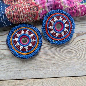 May include: Two round, beaded coasters with a central mirror. The coasters feature concentric circles of blue, orange, red, and white beads. The background includes a patterned textile and a light wood surface.