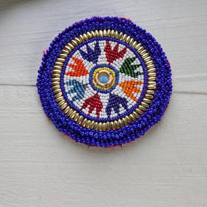 Puede incluir: Un parche circular de cuentas con una piedra azul claro central rodeada por un patrón radial de segmentos rojos, naranjas, verdes y azul oscuro separados por cuentas blancas. El borde consta de cuentas de vidrio en dorado y azul profundo.