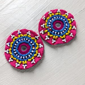 May include: Two circular fuchsia fabric patches featuring intricate beadwork in concentric patterns. The design includes rings of blue, yellow, and white glass beads surrounding a central circular mirror accent, with a sunburst-like beaded edge.