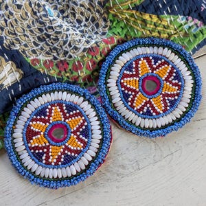 May include: Two circular coasters featuring intricate beadwork in blue, yellow, red, and white. Each coaster has a central circular mirror surrounded by a star-shaped pattern of beads, an outer ring of white oval beads, and a blue beaded border.