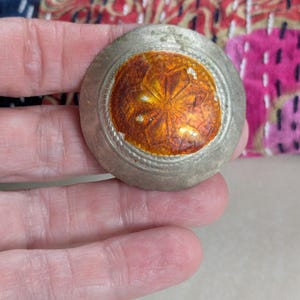 May include: A circular silver-toned metal button featuring a central inlay of translucent orange resin with a stamped floral pattern. The button has a textured border with a beaded edge design surrounding the orange center piece.