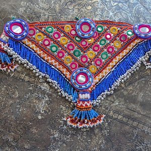 May include: A vibrant, handcrafted textile piece with intricate beadwork and mirror accents. The design features a central triangular pattern in orange, pink, green, and yellow, framed by blue beaded edges and dangling silver-toned embellishments. A unique, colorful accessory.