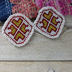 May include: Two square beaded coasters with a red background, yellow and white accents, and a white beaded border. The coasters feature a geometric design and are placed on a wooden surface.