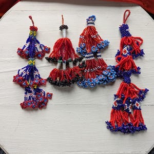 May include: A collection of beaded tassels in red, white, and blue. The tassels are arranged on a white, round surface. The tassels have a variety of shapes and sizes, with intricate beadwork and hanging strands. The color scheme is patriotic.