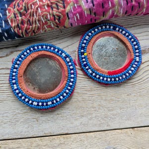 May include: Two round, beaded mirrors with a central reflective surface. The mirrors feature a vibrant design with blue, white, and orange beads. A colorful textile with stitched patterns is visible in the background.