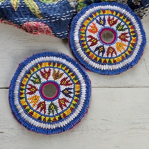 Peut inclure: Deux dessous de verre circulaires ornés de perles aux motifs vifs rouges, jaunes, verts et bleus. Chaque dessous de verre présente un petit miroir central entouré de perles blanches et bleues, formant un design radial détaillé et texturé.