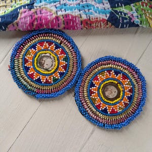 May include: Two round, beaded coasters with intricate patterns. The coasters feature a central design with yellow, red, orange, and white beads, surrounded by gold and blue beads. The background includes a colorful, patterned textile.
