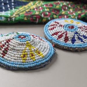 May include: Two round, beaded, decorative pieces with a blue, white, red and yellow colour scheme. The pieces are sewn onto a fabric backing.