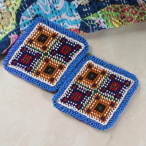 Peut inclure: Deux dessous de verre carrés perlés avec un motif géométrique dans les tons marron, rouge, blanc et vert, encadrés d'une bordure bleue. Les dessous de verre sont posés sur une surface claire, avec un tissu à motifs en arrière-plan.