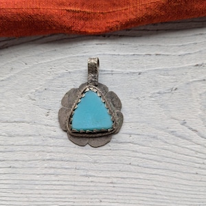 May include: A silver pendant with a turquoise triangle stone set in a floral design. The pendant has a loop at the top for a chain. The background is a white wooden surface with an orange fabric in the upper corner.