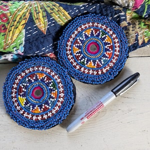 May include: Two circular coasters featuring intricate blue, white, red, yellow, and green beadwork arranged in concentric patterns around a central mirror. A black and silver permanent marker is placed next to the coasters for scale.