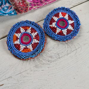 May include: Two circular patches featuring intricate beadwork in shades of blue, red, white, and orange. Each patch has a central circular mirror surrounded by a pink fabric border, with concentric patterns of small glass beads radiating outward.