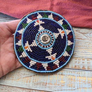 Puede incluir: Un posavasos redondo con cuentas, con un fondo azul oscuro y un botón central plateado. El posavasos presenta un patrón de cuentas blancas, rojas, verdes y naranjas. El posavasos tiene aproximadamente 10 cm de diámetro.