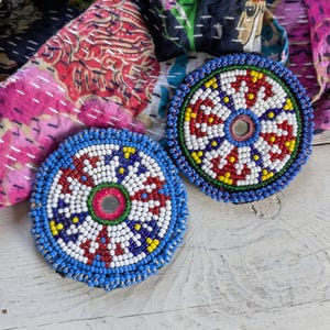 May include: Two circular beaded patches featuring a central mirror surrounded by intricate patterns of white, red, blue, and yellow glass beads. The edges are finished with a border of bright blue beads, creating a vibrant, textured, and symmetrical design.
