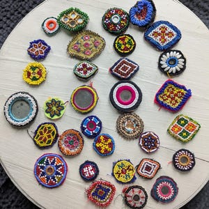 May include: A collection of colorful, beaded and mirrored embellishments in various shapes, including circles, squares, and floral designs. The embellishments feature vibrant colors like red, blue, yellow, and green, with intricate beadwork and reflective mirror accents. They are arranged on a light-colored, round surface.