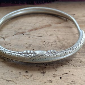 May include: A silver-toned bangle bracelet with a textured, patterned design along the outer edge. The bracelet is circular and appears to be made of metal. The bracelet is resting on a wooden surface.