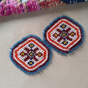 Puede incluir: Dos posavasos con cuentas con un diseño geométrico. Los posavasos presentan una forma de diamante con una combinación de colores rojo, blanco, naranja y azul. Los bordes están forrados con cuentas azules. Los posavasos son probablemente hechos a mano.