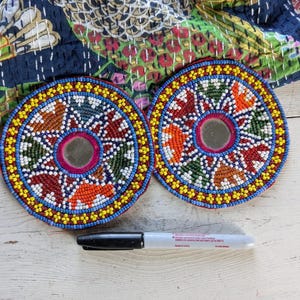 May include: Two circular coasters featuring intricate multi-colored beadwork in red, orange, green, blue, and yellow patterns with a central circular mirror. A black permanent marker is placed below the coasters for scale reference.